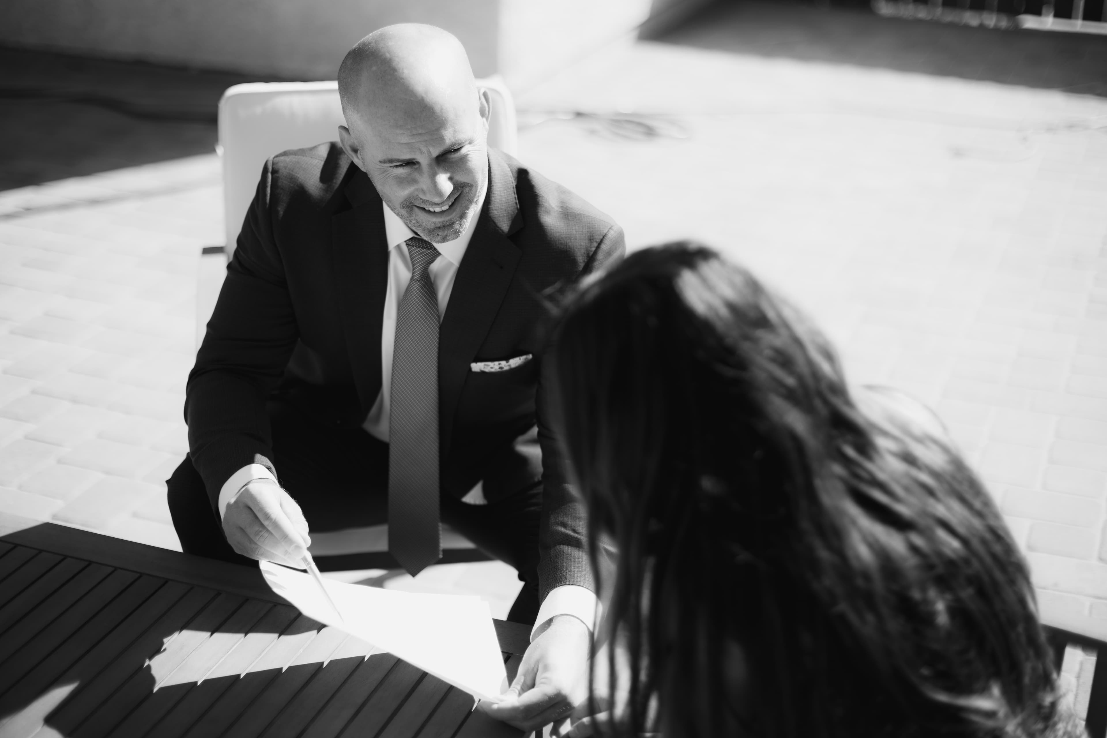 Smiling businessman in a suit reviews a document with a woman at a table outdoors.